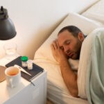 Sick Caucasian man at home self isolating and social distancing in quarantine lockdown during coronavirus covid 19 epidemic, asleep in bed with medication and thermometer on bedside tabl
